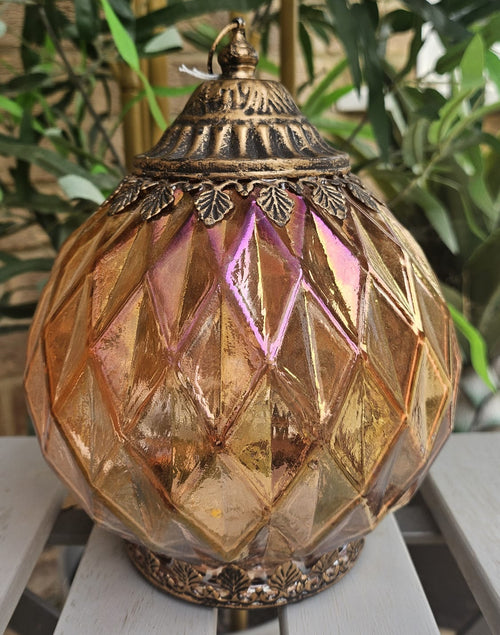 A decorative amber glass dome lamp with a gold-toned metal top and base, featuring a faceted diamond pattern and pinkish reflections, set on a white slatted table with green foliage in the background.