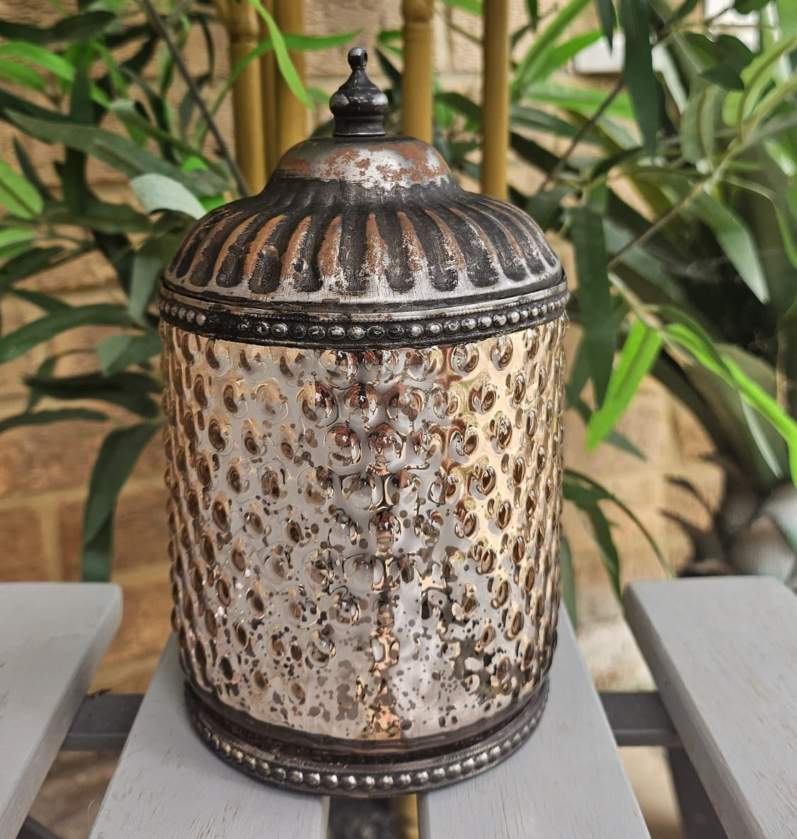 A hammered metal lidded jar with a distressed silver and bronze finish, displayed on a gray wooden table with a background of green plants and bamboo.