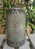 A vintage-style silver metal lidded jar with a textured, diamond-patterned body and ornate filigree metal trim, displayed on a white wooden bench with green plants in the background.