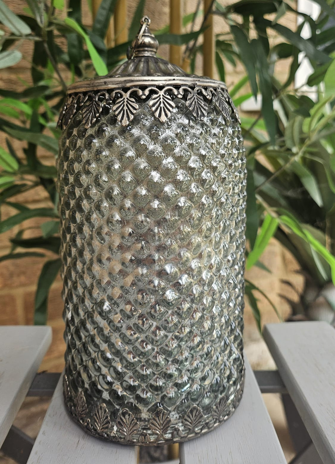 A vintage-style silver metal lidded jar with a textured, diamond-patterned body and ornate filigree metal trim, displayed on a white wooden bench with green plants in the background.